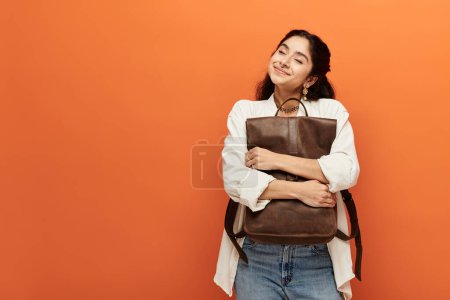 Young indian woman energetically holding backpack against vibrant orange wall.