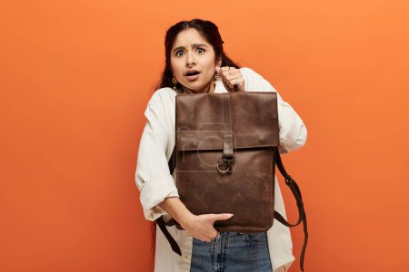 Photo for Young indian woman holding brown backpack against vibrant orange background. - Royalty Free Image
