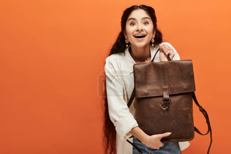 Foto de Mujer india joven mostrando activamente una mochila marrón con estilo contra un fondo naranja brillante. - Imagen libre de derechos