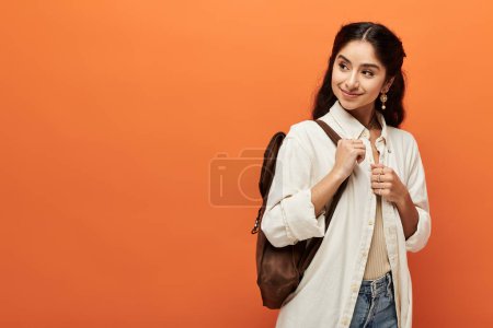 Photo for Young indian woman confidently stands with a backpack against vibrant orange backdrop. - Royalty Free Image