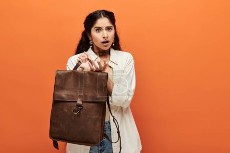 Photo for Young indian woman energetically posing with a brown backpack against vibrant orange background. - Royalty Free Image