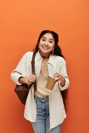 A young indian woman elegantly holding a cup of coffee against an orange wall.