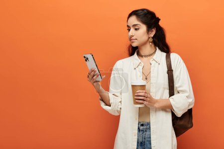 Foto de Una joven india sostiene una taza de café mientras mira la pantalla de su teléfono. - Imagen libre de derechos