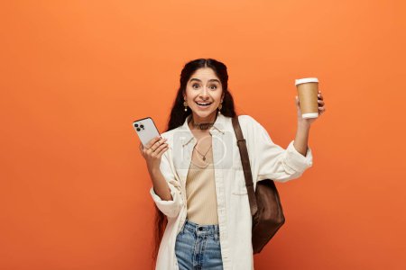 Foto de Una joven india sostiene una taza de café y un teléfono sobre un fondo naranja. - Imagen libre de derechos