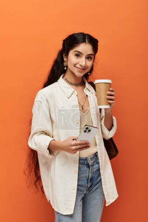 Foto de Una mujer india joven con estilo sostiene alegremente una taza de café sobre un fondo naranja. - Imagen libre de derechos