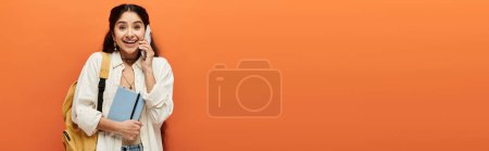 Téléchargez les photos : Une jeune femme indienne avec un sac à dos parle au téléphone sur un fond orange vif. - en image libre de droit