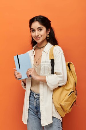 Young indian woman energetically holds a notebook on vibrant orange background.