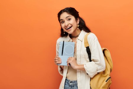 Young indian woman confidently holds notebook on orange background.
