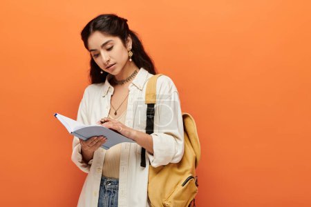 Young indian woman actively writing in notebook against vibrant orange background.