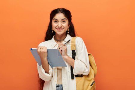Young indian woman energetically holds a notebook against a vibrant orange background.