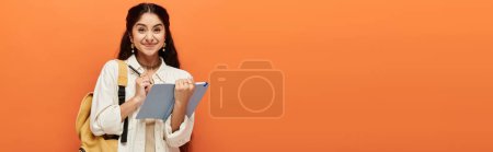 Youthful indian woman holding book against vibrant orange backdrop.