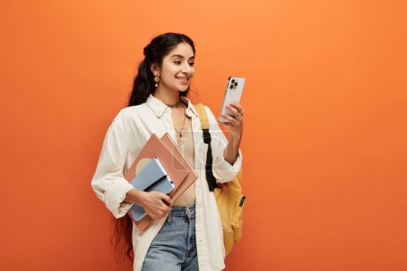 Jeune femme indienne avec sac à dos et téléphone portable sur fond orange.