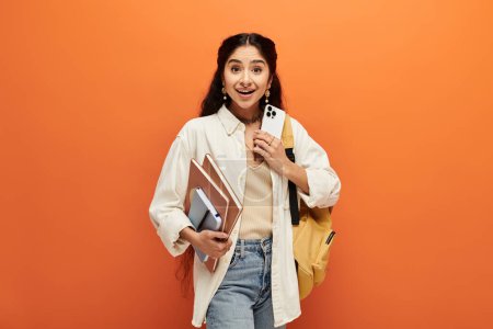 A young indian woman holding a backpack and books against an orange background.