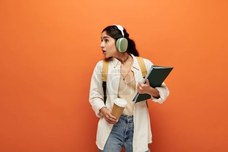 Young indian woman with headphones looks inspired holding notebook against orange background.