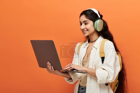 Mujer india joven con auriculares usando portátil sobre fondo naranja.