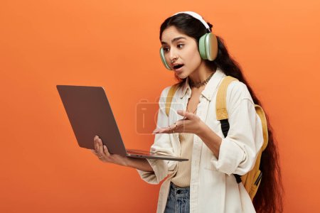 A youthful indian woman immersed in music and work, with headphones and a laptop on an orange background.