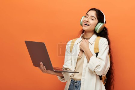 Youthful indian woman wearing headphones, typing on laptop against vibrant orange background.