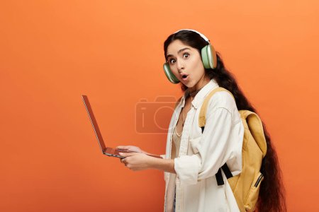 Young indian woman with headphones, using laptop on vibrant orange background.