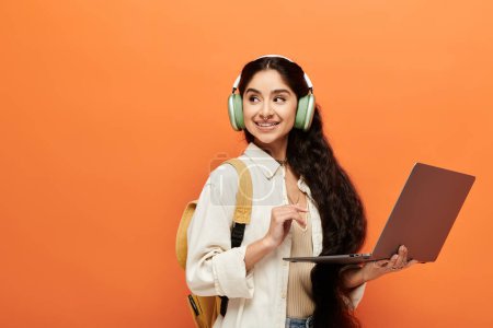 Young indian woman with headphones, holding laptop on orange background.