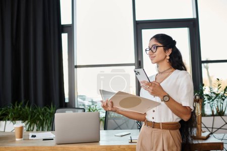 Young indian woman in glasses stands at desk, holding folder.