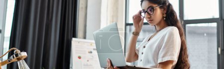 Photo for A indian woman in glasses is using a laptop in an office. - Royalty Free Image