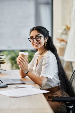 Eine junge indische Frau in Gläsern genießt eine Tasse Kaffee an ihrem Schreibtisch.