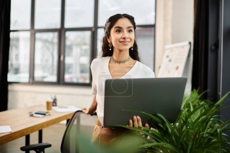 Young indian woman standing in office, using laptop.