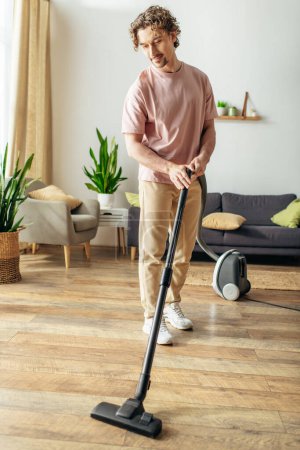 bel homme dans confortable homewear habilement nettoie le sol à l'aide d'un aspirateur.
