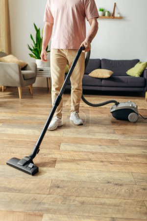 Foto de Un hombre en ropa de casa acogedora utiliza un vacío para limpiar un piso de madera. - Imagen libre de derechos