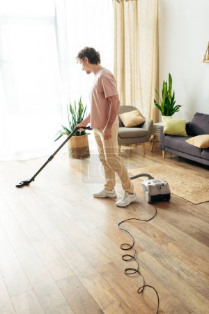 A handsome man in cozy homewear cleans the floor using a vacuum.