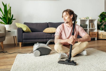 Photo for A handsome man in cozy homewear sitting on the floor while using a vacuum cleaner. - Royalty Free Image
