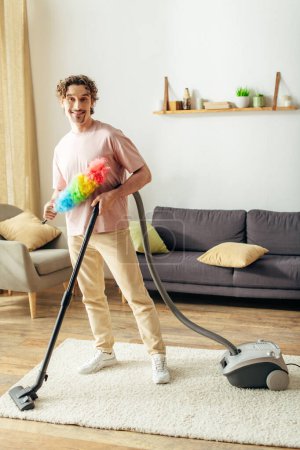 Photo for Handsome man in cozy homewear thoroughly cleans the living room with a vacuum cleaner. - Royalty Free Image