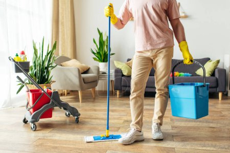 Un hombre guapo en ropa de casa acogedora meticulosamente fregar pisos con un cubo de suministros de limpieza.