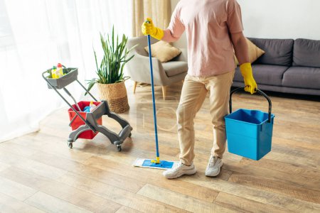 A handsome man in cozy homewear cleans the floor with a mop and bucket.