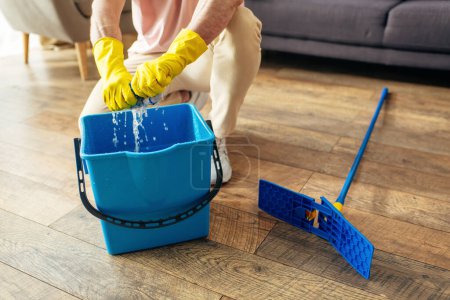 A handsome man in cozy homewear meticulously cleans a blue bucket with yellow gloves.