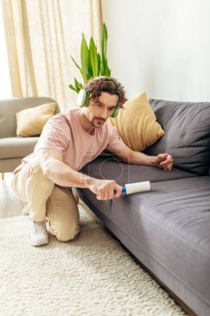 A man in cozy homewear sits on a couch, holding a sticky roller.