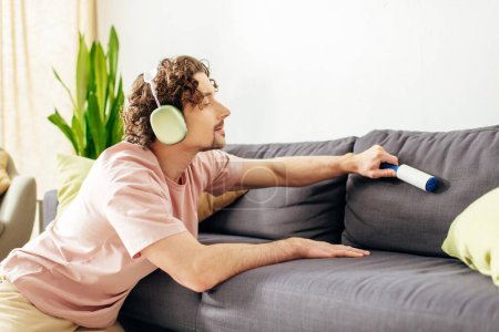 A man in cozy homewear sitting near a couch, listening to music with headphones on.