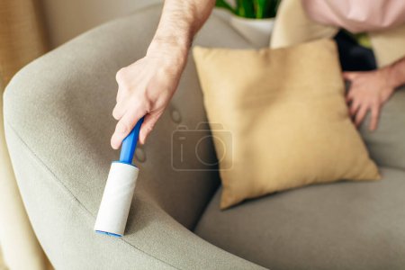 Un hombre en acogedora ropa de casa limpia meticulosamente un sofá usando un rodillo pegajoso azul.
