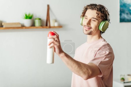 Foto de Joven con auriculares sostiene la botella de aerosol mientras limpia. - Imagen libre de derechos