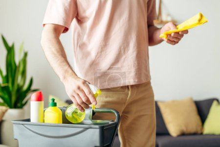 A man in cozy homewear cleans a living room with a yellow sprays.