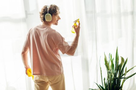 A man in cozy homewear stands in front of a window, listening to music through headphones.