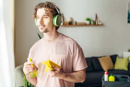 Foto de Un hombre elegante en ropa de casa acogedora escucha música a través de auriculares mientras sostiene un spray. - Imagen libre de derechos