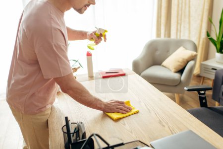 Foto de Hombre guapo en ropa de casa acogedora utiliza una esponja amarilla para limpiar una mesa de madera en una habitación iluminada por el sol. - Imagen libre de derechos