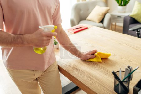 Hombre guapo en ropa de casa acogedora limpieza de una mesa con un paño amarillo.