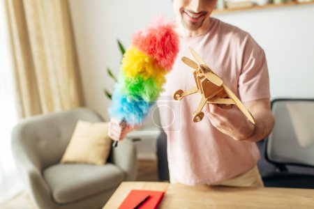 Foto de Hombre guapo en ropa de casa acogedora sosteniendo un avión de juguete y un plumero de color arco iris. - Imagen libre de derechos