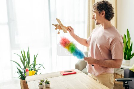 A man in cozy homewear playing with a toy airplane while cleaning at home.