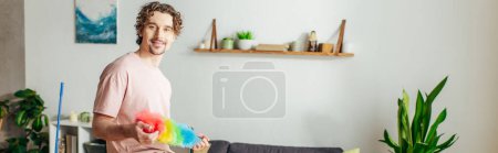 Handsome man in cozy homewear holding a colorful duster in a living room.