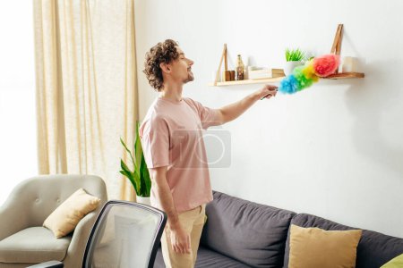 Téléchargez les photos : A handsome man in cozy homewear cleaning a living room with a duster. - en image libre de droit