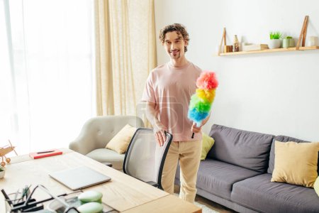 Foto de Hombre guapo en ropa de casa acogedora sosteniendo un plumero vibrante en la elegante sala de estar. - Imagen libre de derechos
