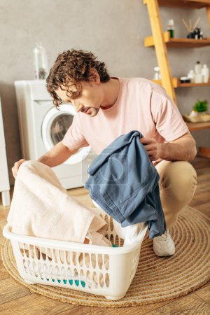 Hombre guapo en ropa de casa acogedora ordenando cuidadosamente la ropa en la cesta de la ropa.